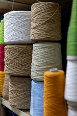 A close-up view of large yarn cones stacked on a shelf in a knitwear factory. The colorful bobbins of white, brown, blue, and orange thread are raw materials for manufacturing.