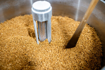 A close-up macro view of a large stainless steel mixing bowl in a confectionery. It is filled with a pile of finely ground nuts or biscuit crumbs, a key ingredient for making chocolate or praline.