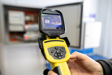 An electrician's hand holds a thermal imaging camera to inspect an open electrical panel, checking for overheating components, hotspots, and potential faults during maintenance.