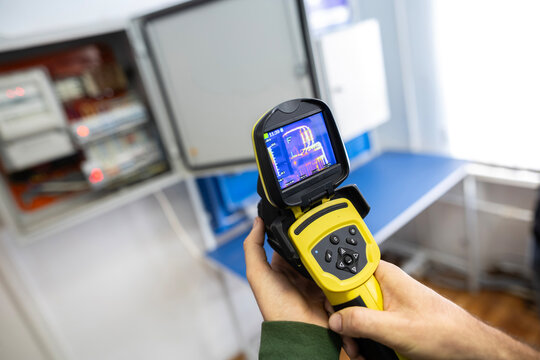 An electrician's hand holds a thermal imaging camera to inspect an open electrical panel, checking for overheating components, hotspots, and potential faults during maintenance.