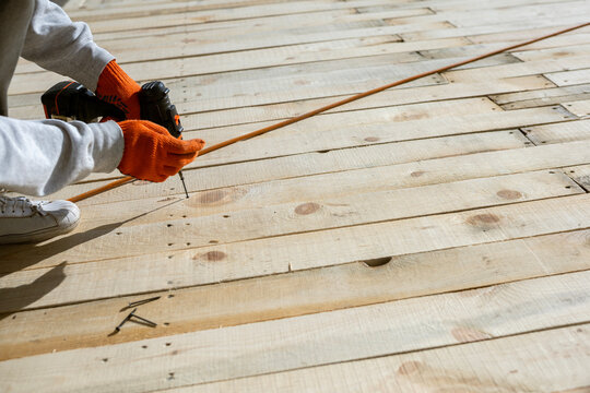 A worker in orange gloves uses a cordless drill to drive a screw, dangerously close to a live extension cord. Unsafe work practice creates a serious risk of electric shock and violates safety rules.