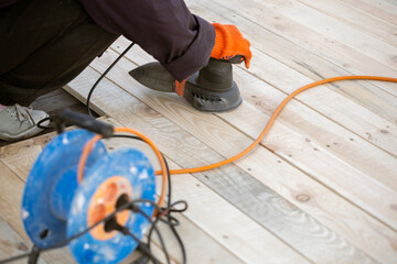 A worker in an orange glove uses an electric orbital sander to smooth a new wooden deck. An orange extension cord and a blue cable reel are on the planks.