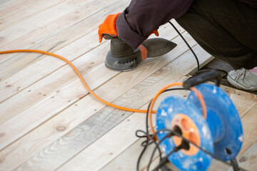 A worker in an orange glove uses an electric orbital sander to smooth a new wooden deck. An orange extension cord and a blue cable reel are on the planks.