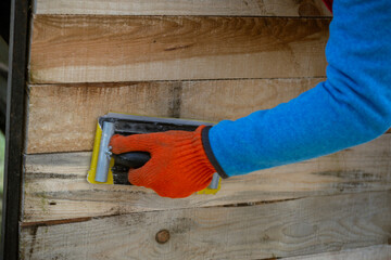 A worker's hand in an orange glove uses a hand sander to smooth wooden planks on a wall. This DIY project involves preparing or refinishing a wood surface with sandpaper.