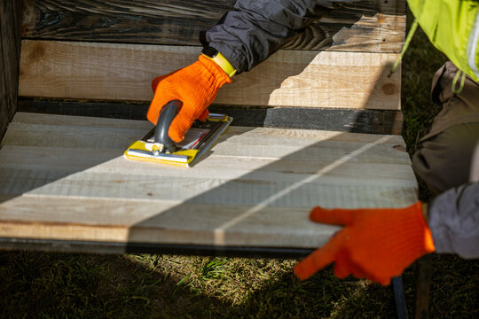 A worker in orange gloves uses a hand sander to smooth the surface of a new wooden bench. This DIY woodworking project involves preparing the planks outdoors in the sun.