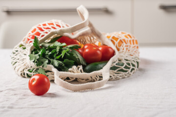 Mesh shopping bag full of fresh vegetables and greens on kitchen table with tablecloth, eco-friendly grocery concept