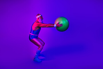 Elderly man in retro athletic jumpsuit pushes a green exercise ball in a neon purple blue studio for fitness fun and vibrant lifestyle imagery
