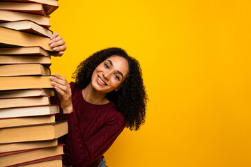 Young mixed race woman smiles while stacking a tall pile of book against a bright yellow background...