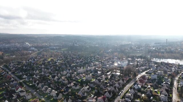 High-angle drone video capturing a residential district of Anykščiai, Lithuania, with compact houses, tree-lined streets, and surrounding forested hills under a hazy autumn sky.