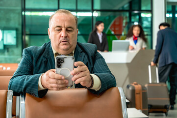 Senior businessman sitting at airport waiting area looking at smartphone and waiting on board