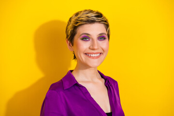 Bright young woman with short blonde hair wearing a purple blouse smiles against a vivid yellow...