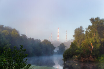 Ethereal morning mist shrouds a tranquil river, contrasting with the looming industrial chimneys. A...