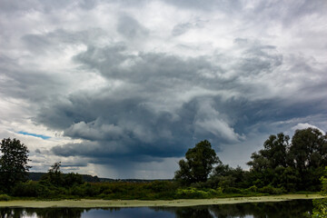 Heavy, brooding storm clouds loom over a tranquil river, reflecting nature's raw power and the impending summer deluge A majestic, dramatic sky scene