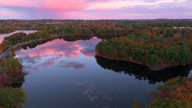 Aerial view of Fisk Pond mirroring the pink and purple sunset sky, surrounded by trees in autumn colors, Natick, Massachusetts, United States.
