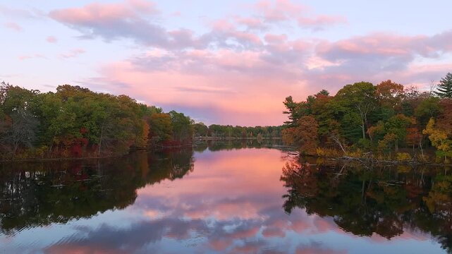 Aerial view of Fisk Pond's still waters mirroring the pastel sky, fringed by trees displaying vibrant autumnal hues, Natick, Massachusetts, United States.
