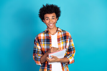 Young man with a plaid shirt holds a notebook and smiles warmly against a bright blue studio background