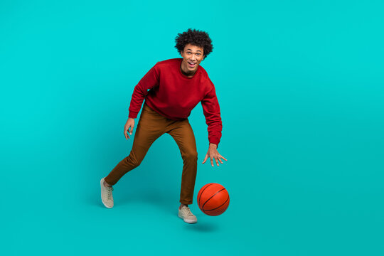 Young male basketball player in red sweater and brown pants dribbles ball against teal background smiling and looking at camera - Powered by Adobe