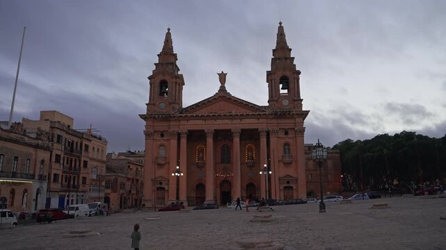 Wide symmetrical view of a Baroque church with twin towers and portico in Valletta, Malta at dusk, pedestrians and parked cars present, soft light and amber lamps.
