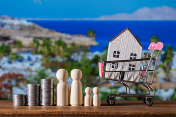 Small wooden toy house in shopping cart, stack of coins and figures of people on nature background. Concept of save money for purchase or construction of home, mortgage and real estate investment