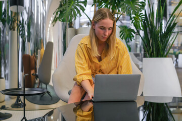 Gen Z freelancer with laptop working in a green coworking space among indoor plants, biophilic design; remote work, focus, productivity, flexible schedule, sustainable work lifestyle.