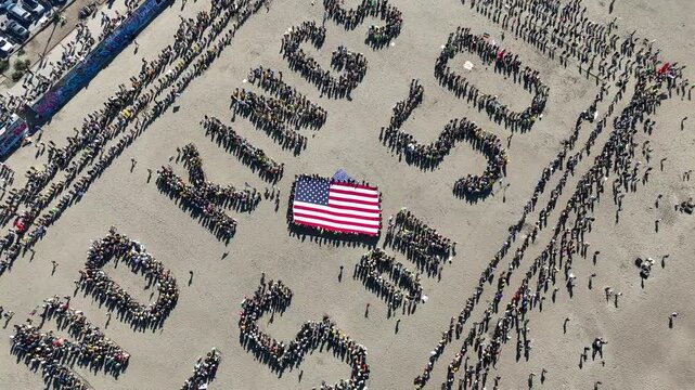 San Francisco, United States - 25 October 2025: Aerial view of crowds forming the words 'NO KINGS YES US 50' around a large American flag on a sandy surface.