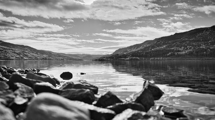 A lake, with stones on the shore in the foreground, mountains and sunbeams.