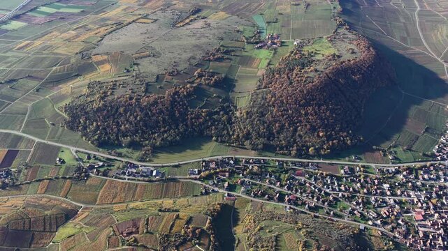 Aerial view of a forested hill casting shadows on the patchwork fields, contrasting with the compact town nestled at its base, Rouffach, Grand Est, France.