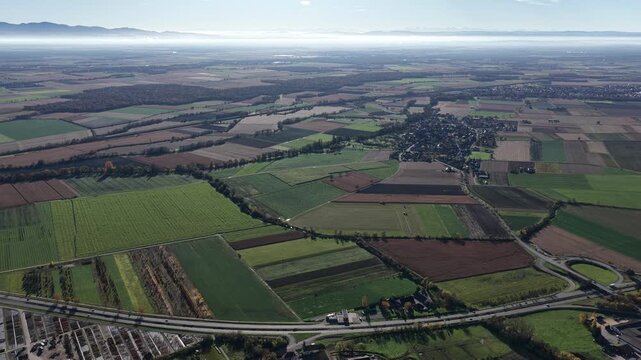 Aerial view of patchwork farmlands and a distant town nestle under a hazy sky, bisected by roads, creating a blend of rural and urban textures, Rouffach, Grand Est, France.