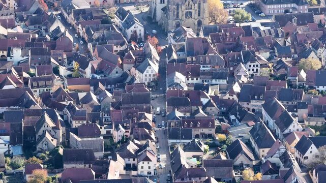 Aerial view of tightly packed buildings and a church tower, the red rooftops contrasting with the autumn trees, Rouffach, Grand Est, France.