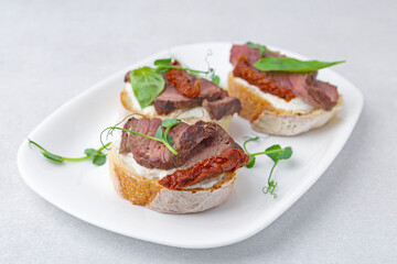 Bruschettas with roast beef, cream cheese, basil and sun-dried tomatoes on light grey table, closeup