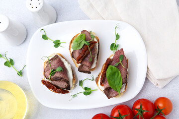 Bruschettas with roast beef, cream cheese, basil, sun-dried tomatoes and fresh vegetables on light grey table, flat lay