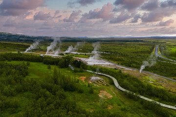 Aerial scene shows steam plumes, hot pools, and a curving boardwalk at the Geysir Geothermal Area in Haukadalur, Iceland, under soft evening light and pastel clouds.