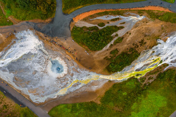 Top down aerial shows blue hot spring, mineral streaks, white and yellow terraces, winding paths and roads, and visitors on boardwalks in daytime diffuse light.
