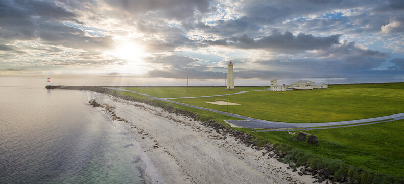 Aerial view shows a white lighthouse and low buildings on a grassy Iceland peninsula at golden hour, pale sand and dark rocks, a breakwater beacon, and calm reflective sea.