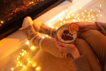 Woman with delicious hot drink and Christmas lights near fireplace at home, closeup