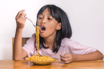 Hungry little girl eating noodles with a fork while sitting at table