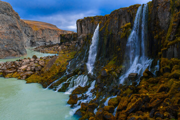 Water pours over dark basalt into turquoise water, mossy rocks and rugged canyon walls frame the view in Iceland. Soft overcast light enhances texture and contrast.