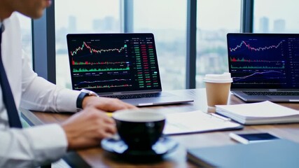 A businessman at a desk with two laptops displaying trading charts, a coffee cup, and documents - Powered by Adobe