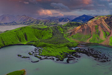 Aerial scene in Landmannalaugar, Iceland shows moss green mountains, dark basalt ridges, lava islets, braided channels, and crater rims at evening with pink light.