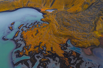 Naklejka premium Aerial view of milky turquoise glacial water winding through dark channels on a rust orange delta in Iceland near Landmannalaugar, under soft diffused autumn light.