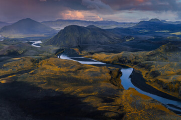 Aerial view shows a winding glacial river across dark volcanic plains and mossy ridges near Landmannalaugar, Iceland, in soft evening light under storm clouds.