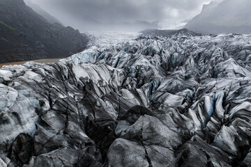 Aerial view shows a jagged glacier in Vatnajokull National Park, Iceland, crevassed ice with dark ash, distant mountains, and a milky glacial lagoon at left in cool light. © Aerial Film Studio