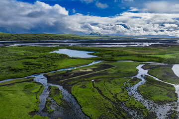 Aerial view of an Iceland river delta near Landmannalaugar, braided channels and lagoons, volcanic ridges and distant snowy peaks, emerald moss and black sands.