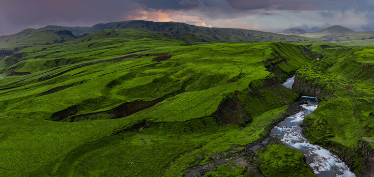 Aerial view of Iceland highlands at dusk, with bright green lava field, narrow river in a rugged gorge, volcanic plateaus, dark basalt, and storm clouds with sunset light. - Powered by Adobe