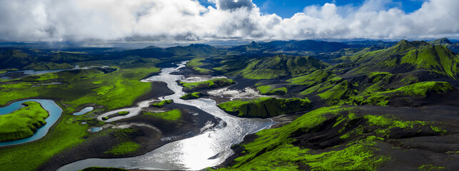 Aerial panorama shows a braided glacial river through basalt and ash in Iceland, with moss green slopes, rhyolite mountains, and midday light under patchy clouds.