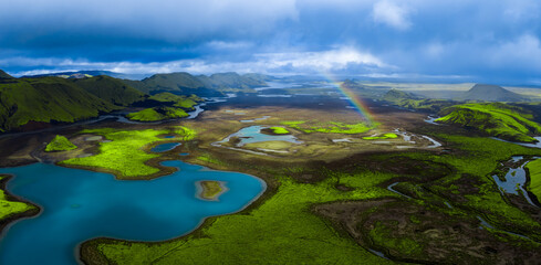 Aerial view of Landmannalaugar in Fjallabak Nature Reserve, rainbow arches above mossy lava, crater plains, braided rivers, sapphire lakes, and dark volcanic ridges. © Aerial Film Studio
