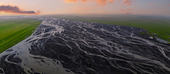 Braided silvery rivers fan across a black sandur near Vik, Iceland, in evening summer light. Channels snake over volcanic ash, framed by green farms and long shadows.