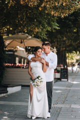 Naklejka premium Wedding photography, portrait of happy smiling newlyweds, stylish groom and beautiful young brunette bride, hugging outdoors on the street in the city, in the park.