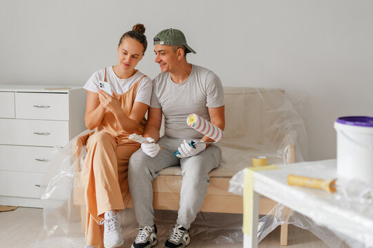 Two happy individuals sit on a couch surrounded by painting supplies, looking at their phones and enjoying their time together while preparing to redecorate