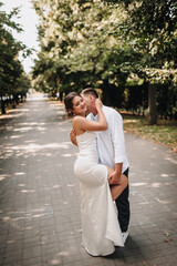Naklejka premium Wedding photography, portrait of happy smiling newlyweds, a young groom and a beautiful sweet brunette bride, tenderly hugging outdoors on a street in the city, in a park.